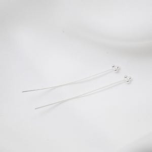 Könnte beinhalten: Ein Paar silberne Ohrringe. Jeder Ohrring hat einen langen, dünnen Draht mit einer Ansammlung von drei kleinen, runden Perlen an der Spitze. Die Ohrringe sind vor einem weißen Hintergrund platziert, was ihr minimalistisches Design hervorhebt.