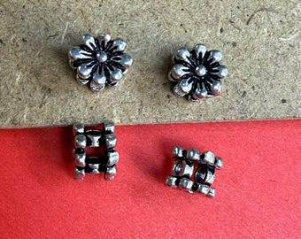 May include: Four small, silver-colored metal beads. Two are flower-shaped with detailed petals, and two are rectangular with a grid-like design. The beads are on a red and tan surface.
