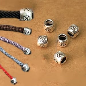 Pode incluir: Contas de metal prateado com um design repetitivo em forma de trevo. Várias contas são mostradas, algumas presas a cordões coloridos. As contas têm uma forma cilíndrica com um buraco no centro.