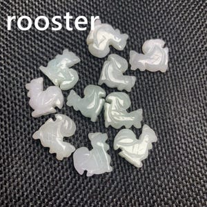 Pode incluir: Uma cole&ccedil;&atilde;o de pequenas figuras de galo esculpidas em tons de branco e verde p&aacute;lido. A palavra "rooster" est&aacute; impressa em branco no topo. As figuras est&atilde;o dispostas sobre uma superf&iacute;cie escura e texturizada.