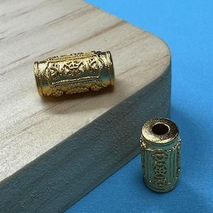 Puede incluir: Dos cuentas de metal de color dorado con diseños intrincados. Las cuentas son cilíndricas y tienen un acabado liso y pulido.