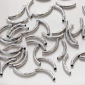 Pode incluir: Um grupo de contas de conexão de metal prateadas com uma superfície texturizada. As contas têm a forma de um semicírculo.