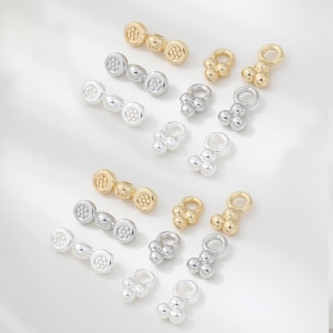 May include: An assortment of small jewelry findings in gold and silver tones. The findings include connector bars with circular designs and charm pendants with three-sphere clusters. The pieces are arranged on a white surface.