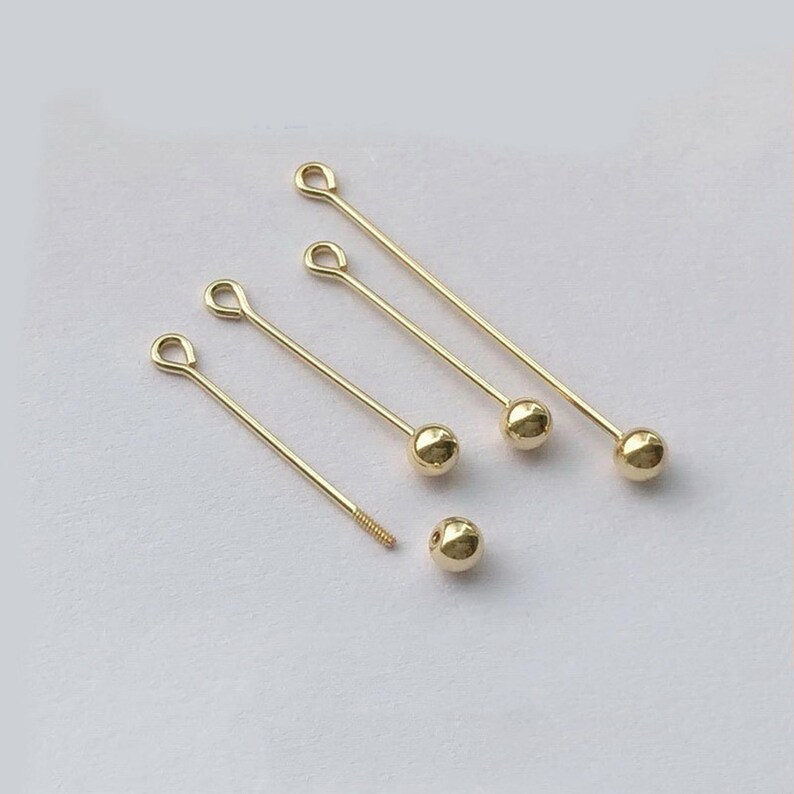 Peut inclure: Des &eacute;l&eacute;ments de bijouterie dor&eacute;s, comprenant plusieurs &eacute;pingles droites avec des boucles et des accents en forme de boule. Une &eacute;pingle a une extr&eacute;mit&eacute; filet&eacute;e. Une perle en forme de boule est &eacute;galement pr&eacute;sente. Les &eacute;l&eacute;ments sont dispos&eacute;s sur une surface blanche.