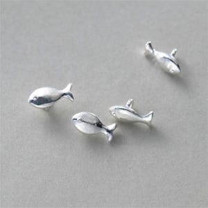 May include: Four small, silver-colored fish figurines are scattered on a gray surface. Each fish has a rounded body, a tail fin, and a small dorsal fin. The figurines appear to be made of metal and have a polished finish.