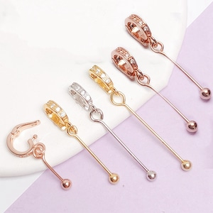 May include: An assortment of dangle charm connectors in various metallic finishes, including gold, silver, and rose gold. Each connector features a small, decorative hoop adorned with clear gemstones and a long, slender bar ending in a small ball.