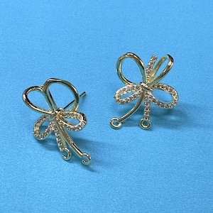 Peut inclure: Une paire de supports de boucles d'oreilles dorés avec un motif de nœud et des strass transparents. Les supports ont un poteau et une boucle pour attacher un charme suspendu.