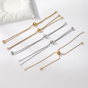 May include: Five adjustable slider bracelets featuring thin snake chains. Three bracelets are gold-toned and two are silver-toned. Each bracelet includes a central sliding bead mechanism and small spherical end caps on the chain terminals.