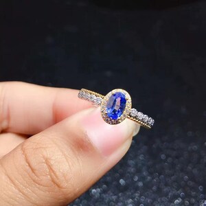 Op de afbeelding: Een gouden ring met een centrale ovaal geslepen blauwe edelsteen omringd door een halo van kleine witte steentjes. De ringband is aan beide zijden bezet met een rij kleine witte steentjes, wat zorgt voor een gedetailleerde uitstraling.