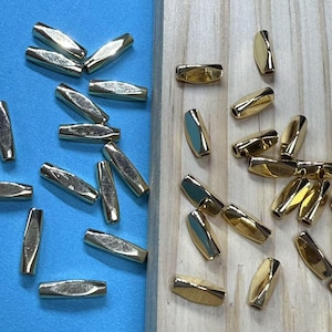 Puede incluir: Primer plano de dos montones de cuentas de metal de color dorado. Las cuentas son de forma ovalada y tienen un diseño facetado. Las cuentas están dispuestas sobre una superficie azul y una superficie de madera.
