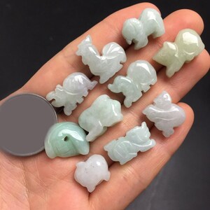 Pode incluir: Uma cole&ccedil;&atilde;o de pequenas figuras de animais em jade esculpido, incluindo um galo, um porco e um c&atilde;o. As figuras s&atilde;o verde claro e brancas, com um acabamento liso e polido. A cole&ccedil;&atilde;o &eacute; segurada em uma m&atilde;o.