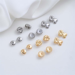 May include: An assortment of earring backs in silver and gold tones. The backs include various styles, such as flower-shaped and heart-shaped designs. The earring backs are displayed on a white surface.