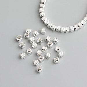 May include: A scattering of silver square beads with a hole in the centre. The beads are arranged in a random pattern on a grey surface. A silver chain of square beads is visible in the background.