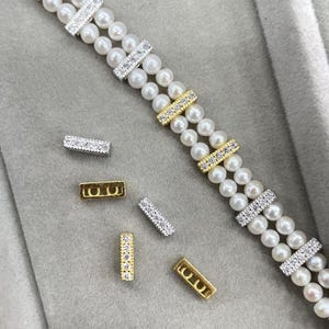 Può includere: Un bracciale di perle a doppio filo con distanziatori rettangolari alternati color argento e oro, incastonati con cristalli trasparenti. Diversi distanziatori rettangolari sciolti, alcuni color oro e altri color argento con cristalli, sono posizionati accanto.