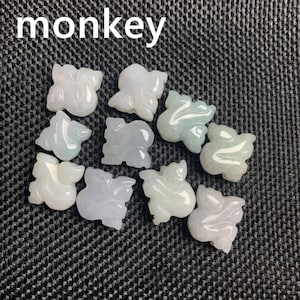 Pode incluir: Uma cole&ccedil;&atilde;o de pequenas figuras de macaco de jade esculpidas em v&aacute;rios tons de branco, verde e cinza. A palavra "monkey" est&aacute; impressa em branco no topo. As figuras est&atilde;o dispostas sobre uma superf&iacute;cie texturizada cinza escuro.
