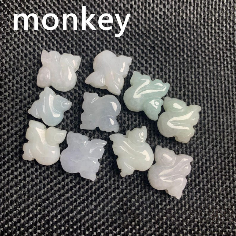 Pode incluir: Uma cole&ccedil;&atilde;o de pequenas figuras de macaco de jade esculpidas em v&aacute;rios tons de branco, verde e cinza. A palavra "monkey" est&aacute; impressa em branco no topo. As figuras est&atilde;o dispostas sobre uma superf&iacute;cie texturizada cinza escuro.