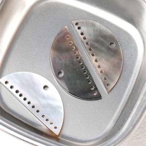 Puede incluir: Tres componentes de joyería iridiscentes en forma de media luna con agujeros perforados, descansando en una bandeja plateada. Dos son de color gris oscuro y uno blanco. Los componentes tienen un brillo nacarado, lo que sugiere que están hechos de nácar.