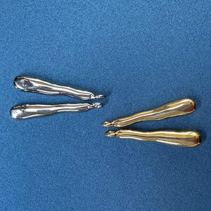 Op de afbeelding: Twee paar oorbellen, één paar is zilver en het andere is goud. Beide paren zijn lang en gebogen.