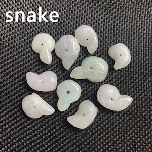Pode incluir: Uma cole&ccedil;&atilde;o de nove pingentes de cobra de jade esculpida em tons de branco e verde p&aacute;lido. Cada pingente tem um furo para enfiar e &eacute; exibido em uma superf&iacute;cie texturizada cinza escuro. A palavra "snake" est&aacute; escrita em branco.