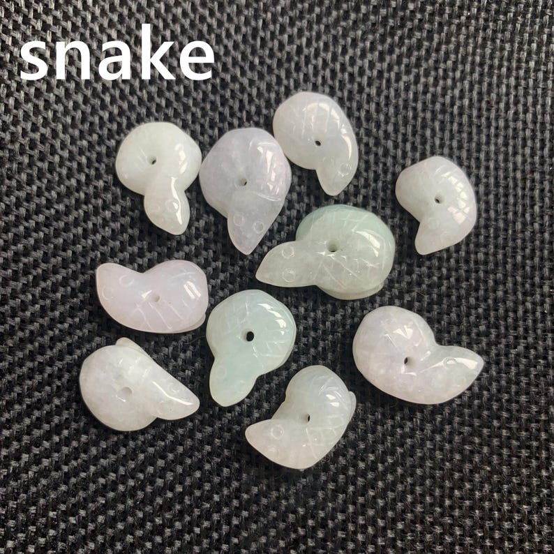Pode incluir: Uma cole&ccedil;&atilde;o de nove pingentes de cobra de jade esculpida em tons de branco e verde p&aacute;lido. Cada pingente tem um furo para enfiar e &eacute; exibido em uma superf&iacute;cie texturizada cinza escuro. A palavra "snake" est&aacute; escrita em branco.