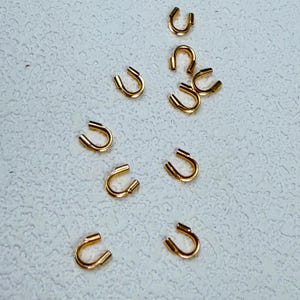 Puede incluir: Una colección de pequeños hallazgos de joyería dorados en forma de U, dispersos sobre una superficie texturizada de color blanquecino. Estos hallazgos parecen ser de extremo abierto, posiblemente para adjuntar a otros componentes de joyería.
