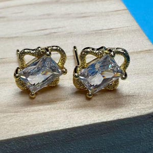 Puede incluir: Un par de pendientes de botón dorados. Cada pendiente presenta una piedra preciosa rectangular y transparente engastada en un marco decorativo de color dorado. Los pendientes se exhiben sobre una superficie de madera de color claro.