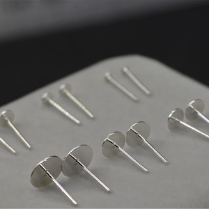 Peut inclure: Un ensemble de tiges d'oreille en argent avec des sommets ronds. Les tiges sont dispos&eacute;es en rang&eacute;e sur une surface blanche.