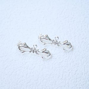 May include: Two silver-colored jewelry clasps. Each clasp features a decorative design with a central, ridged bar and ornate ends. The clasps are displayed on a textured, white surface.
