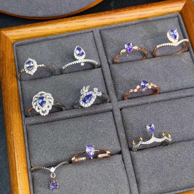 May include: A wooden box with compartments containing nine silver and gold rings with purple gemstones. The rings are set with various designs, including a heart, a crown, and a teardrop.