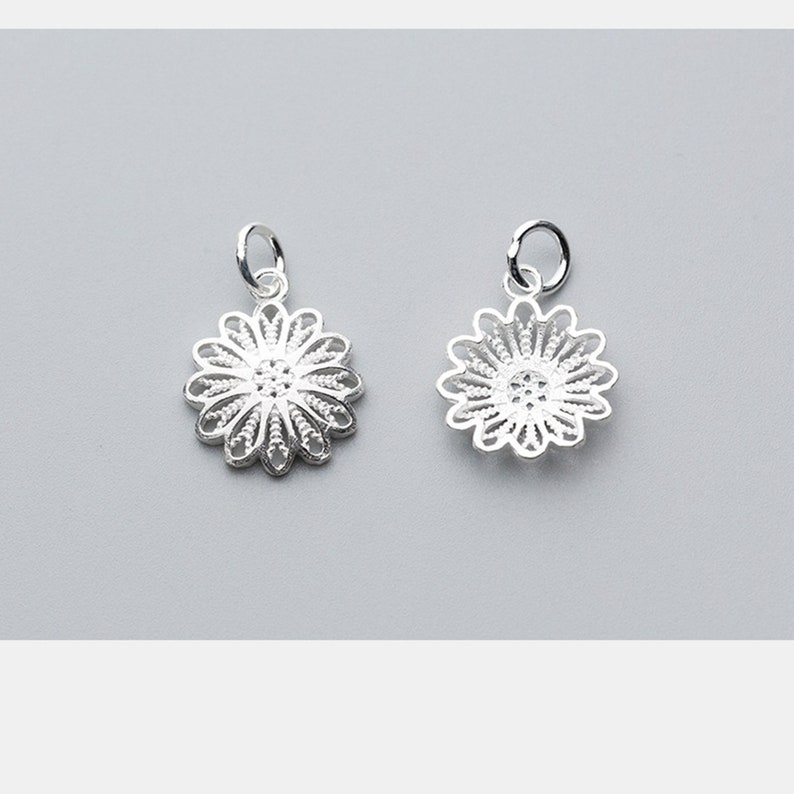 May include: Two silver charm pendants in the shape of a delicate, intricate flower with a circular center. The charms have a loop at the top for attaching to a necklace or bracelet.