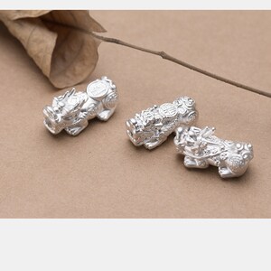 Op de afbeelding: Drie zilveren Chinese drakencharms met ingewikkelde details. De charms zijn in de vorm van een draak met een munt op zijn rug. De charms liggen op een lichtbruin oppervlak.