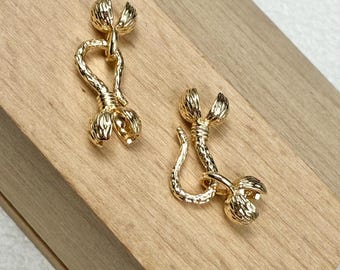 May include: Two gold-colored, ornate clasps. Each clasp features a textured, floral-like design with a hook and loop closure. The clasps are displayed on a light brown wooden surface.