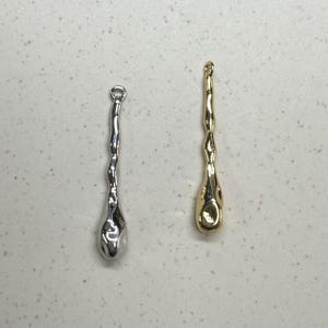 Op de afbeelding: Twee metalen bedels in de vorm van een traan, een zilveren en een gouden. De bedels hebben een getextureerd oppervlak en een lus aan de bovenkant om aan een ketting of armband te bevestigen.
