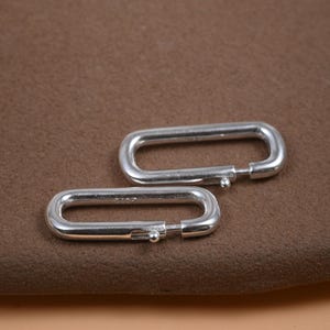 May include: Two silver-colored, rectangular carabiner clasps. Each clasp has a hinged opening with a small ball closure. The clasps are displayed on a brown surface.