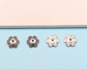 May include: Three silver flower-shaped metal beads with a hole in the center. The beads are on a light blue surface.