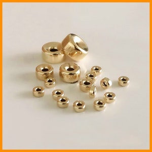 May include: A collection of gold-toned metal beads of various sizes. The beads are round with a hole in the center. They are arranged on a white surface.