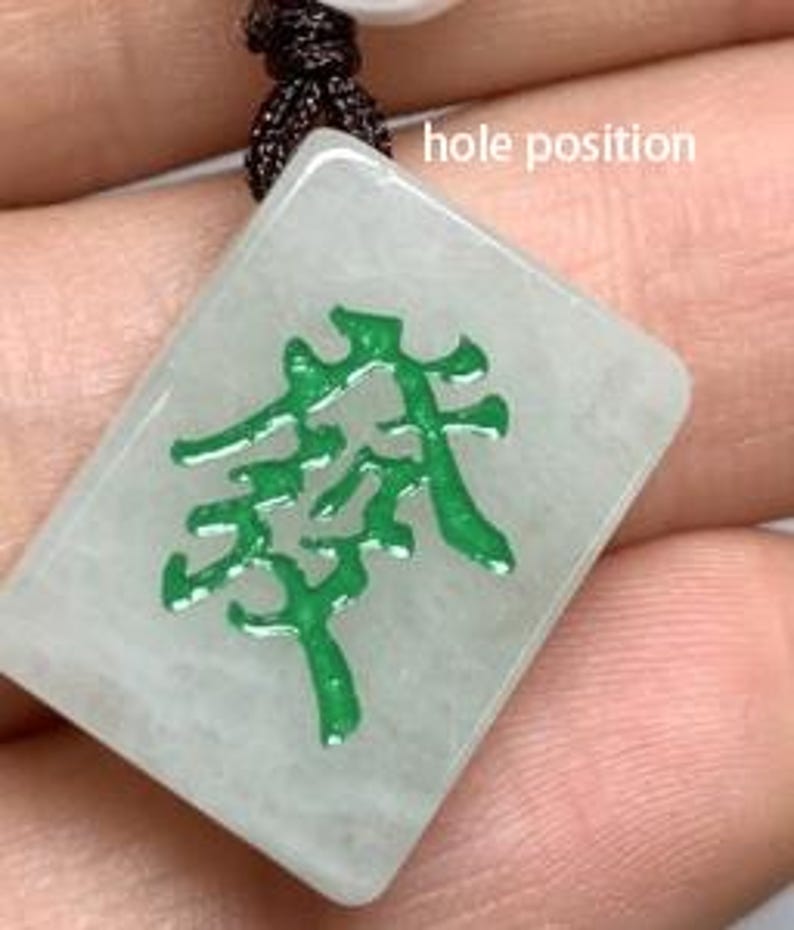 May include: A green jade mahjong tile pendant with a white background and a green inscription. The tile has a hole at the top for a cord or chain.