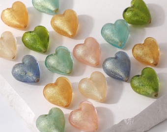May include: A collection of heart-shaped beads in various colors, including gold, green, blue, and pink. The beads have a glittery, translucent appearance and are scattered on a white surface. These decorative beads are suitable for jewelry making or craft projects.