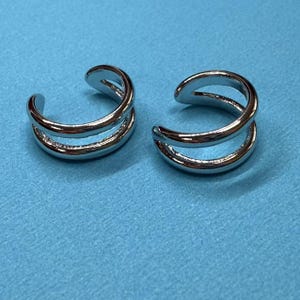 May include: Two silver-colored ear cuffs. Each cuff has a double-band design, with a smooth, polished finish. The ear cuffs are displayed on a light blue surface, creating a contrast with the shiny metal.