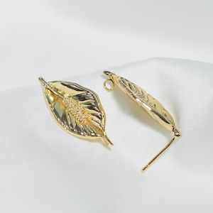 Puede incluir: Dos pendientes dorados con forma de hoja. Los pendientes tienen un patrón de vetas detallado y un centro texturizado. Un pendiente tiene un poste para perforar y el otro tiene un bucle. Sobre un fondo blanco.