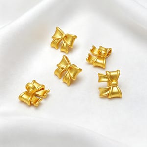Op de afbeelding: Vijf kleine, matgoudkleurige strikbedels, elk voorzien van een centrale knoop en twee lussen. Deze metalen strikvormige onderdelen zijn ontworpen met een klein oogje aan de bovenkant voor bevestiging en hebben een gladde, gelijkmatige afwerking.