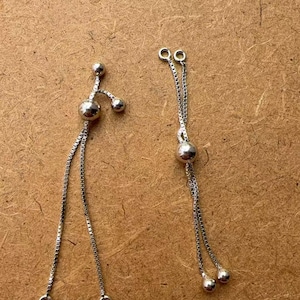 May include: Two silver-toned chain earrings with a delicate design. Each earring features a small, round charm and a series of dangling spheres. The earrings are displayed against a neutral, textured background.