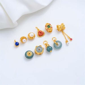May include: A collection of gold-toned jewelry charms featuring various designs, including a blue lapis lazuli stone, white pearls, a red floral-shaped stone, a green rectangular stone, a jade ring, and a dangling charm with a pearl and red bead.