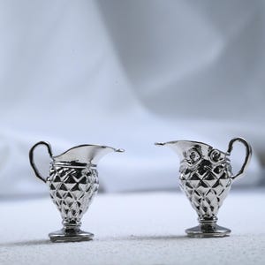 Op de afbeelding: Twee kleine, zilverkleurige roomkannetjes. Elk kannetje heeft een handvat, een tuit en een diamantvormig lichaam. De kannetjes worden gepresenteerd tegen een zachte, witte achtergrond.