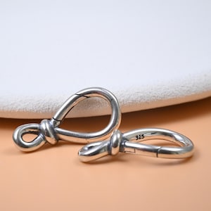 Puede incluir: Dos cierres plateados en forma de nudo. Un cierre está abierto y el otro está cerrado. Los cierres tienen un acabado pulido y un diseño simple y elegante. El número "925" está estampado en uno de los cierres.