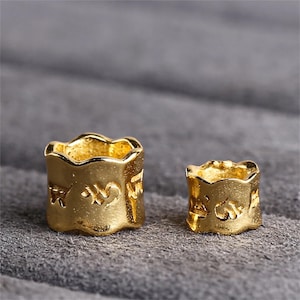 Puede incluir: Dos anillos de metal de color dorado con diseños intrincados y letras en relieve. Los anillos son de diferentes tamaños y tienen un borde ondulado.