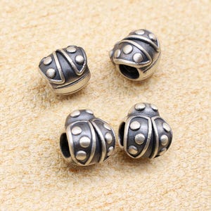 Peut inclure: Quatre petites perles argentées en forme de coccinelle. Chaque perle a une finition sombre oxydée avec des détails circulaires en relief. Les perles sont conçues avec un trou pour l'enfilage, adaptées à la fabrication de bijoux ou à l'artisanat.