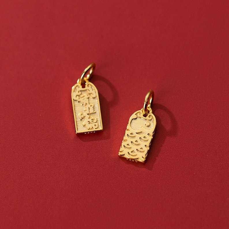 May include: Two gold charms, one with Chinese characters and the other with a fish design. The charms are on a red background.