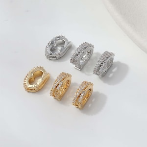 May include: An assortment of jewelry findings, including gold and silver-toned earring clasps and decorative hoops. The clasps have a heart shape, while the hoops are adorned with small, sparkling stones.