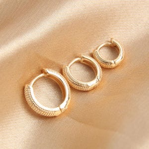 Puede incluir: Tres pendientes de aro dorados de diferentes tama&ntilde;os, colocados sobre una superficie satinada. Los pendientes tienen un dise&ntilde;o texturizado y punteado. El pendiente m&aacute;s peque&ntilde;o est&aacute; en la parte superior derecha, el m&aacute;s grande en la parte inferior izquierda.